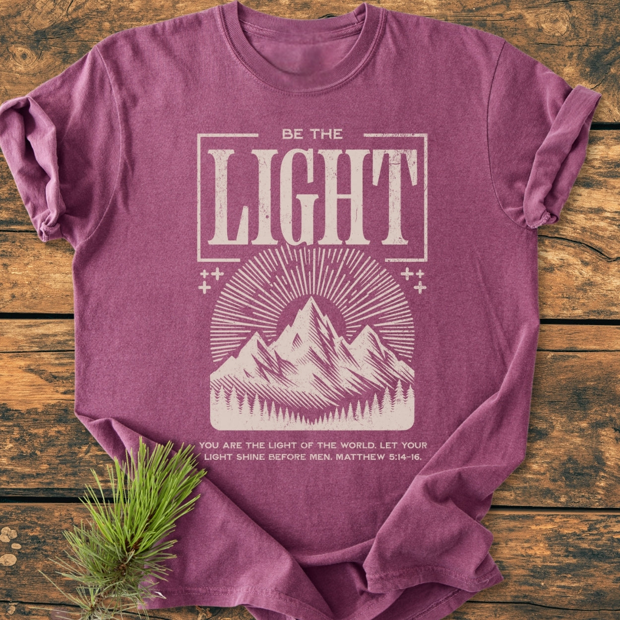 Be The Light - Vintage Wash Tee featuring a mountain illustration with sunrays and a Bible verse, displayed on rustic wood with pine sprig. Made of soft, durable ring-spun cotton.