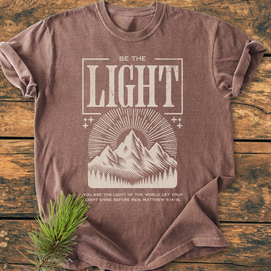 Be The Light - Vintage Wash Tee features a mountain and sunbeam graphic with an inspiring Bible verse on a brown T-shirt, highlighting its relaxed fit and durable Comfort Colors garment-dyed fabric.