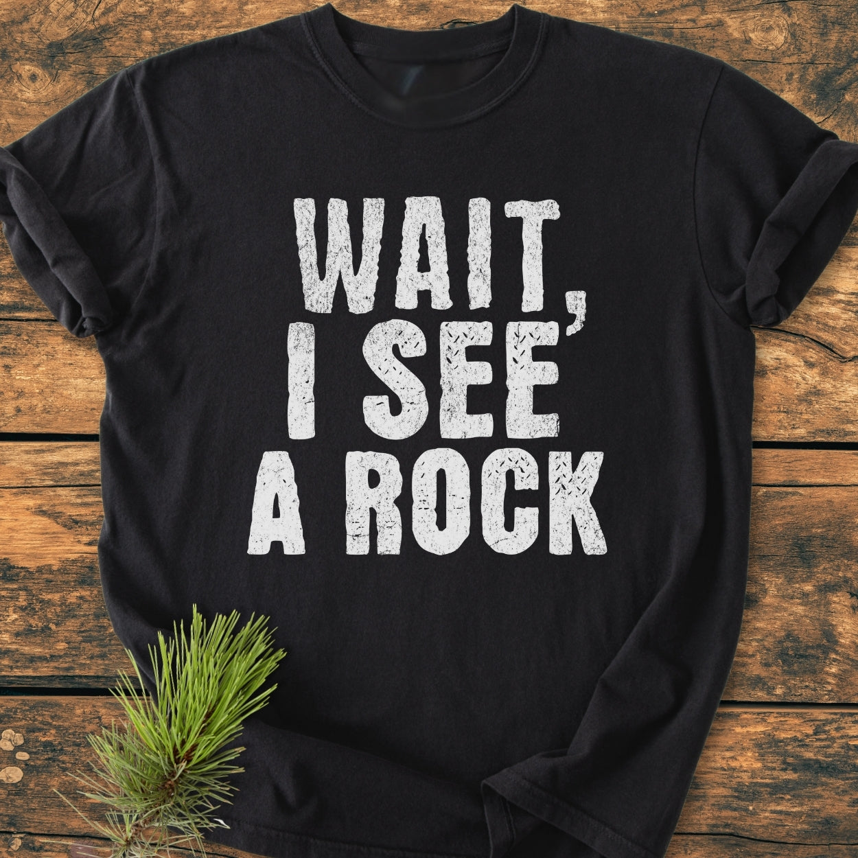 Wait I See A Rock - Vintage Wash Tee displayed on wood, featuring distressed white text. A pine sprig adds a natural touch. Made from soft, garment-dyed cotton for comfort and durability.
