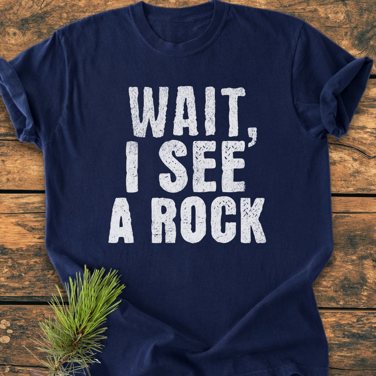 Navy-blue Wait I See A Rock - Vintage Wash Tee with distressed white lettering, laid flat on wood. Accented with a pine sprig, showcasing its relaxed fit and soft, garment-dyed cotton fabric.