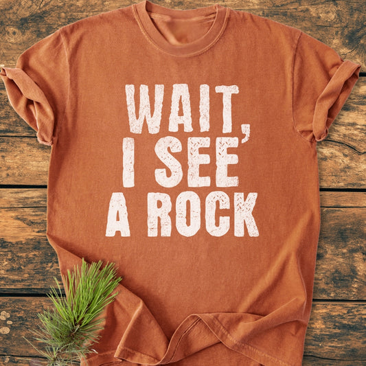 The Wait I See A Rock - Vintage Wash Tee features a bold, distressed white print on a rust-orange T-shirt, laid flat on a wooden surface with a small pine sprig.