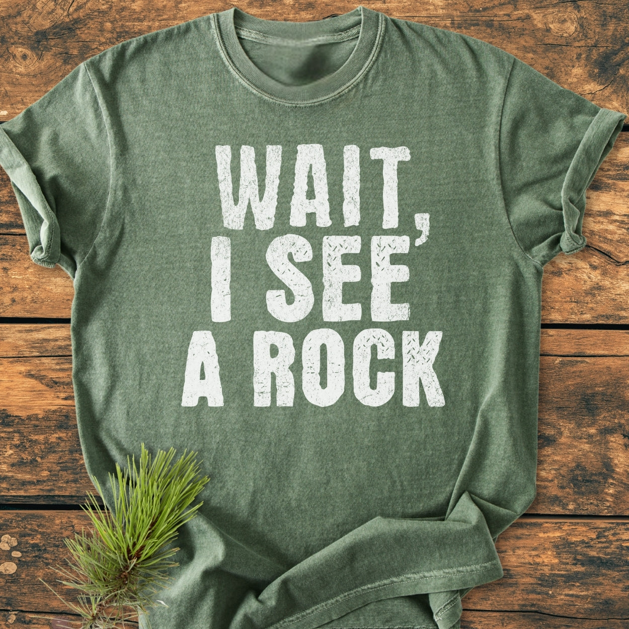 Heather-green Wait I See A Rock - Vintage Wash Tee on rustic wood, featuring distressed white text and a pine sprig beneath. Crew-neck, relaxed fit, double-stitched, garment-dyed for softness and durability.