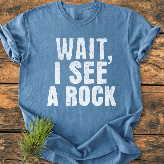 Heather-blue Wait I See A Rock - Vintage Wash Tee laid flat, featuring distressed white text. Pine branch accentuates the soft, relaxed fit and durable garment-dyed fabric.