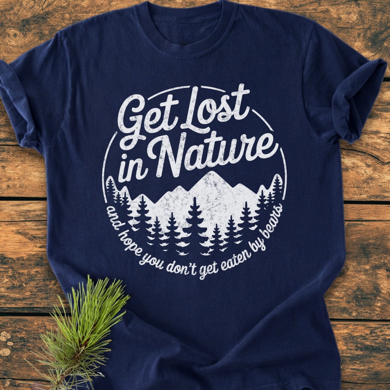 Get Lost In Nature - Vintage Wash Tee: Navy-blue T-shirt with distressed white mountain and pine graphic, bold script slogan, laid on wood. Features durable, soft-washed 100% cotton, relaxed fit, ideal for everyday wear.