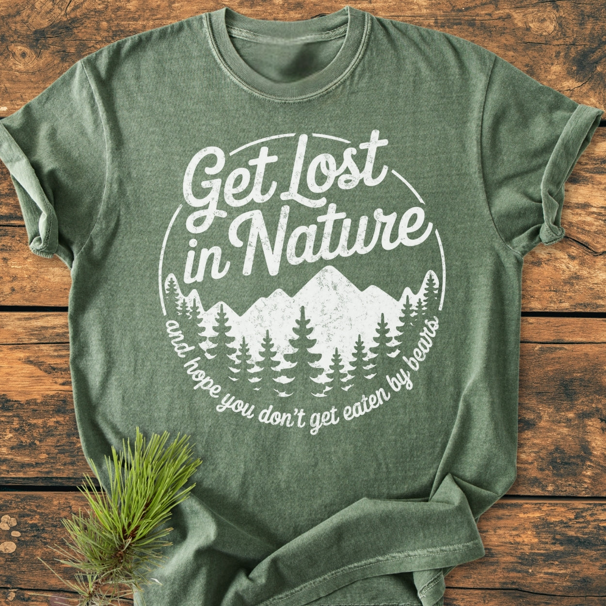 Get Lost In Nature - Vintage Wash Tee: Forest-green T-shirt with playful mountain and pine tree graphic, laid flat on wood. Features durable, soft, garment-dyed 100% ring-spun cotton, offering relaxed fit.