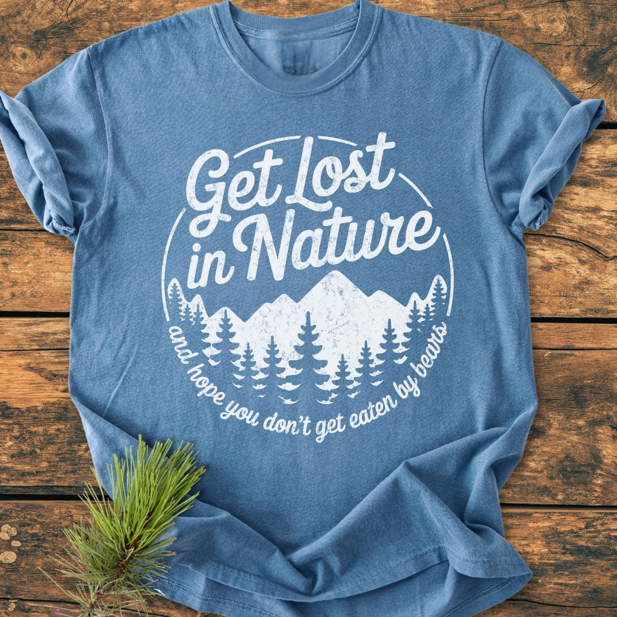 Get Lost In Nature - Vintage Wash Tee features a heather-blue hue with a white circular graphic of mountains and trees, humorously captioned. Laid on wood, a pine sprig complements the scene.