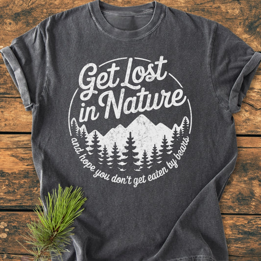 Get Lost In Nature - Vintage Wash Tee: A dark-gray, short-sleeve T-shirt featuring a white mountain and pine tree graphic with humorous text, laid on a wooden surface with a pine branch beside it.
