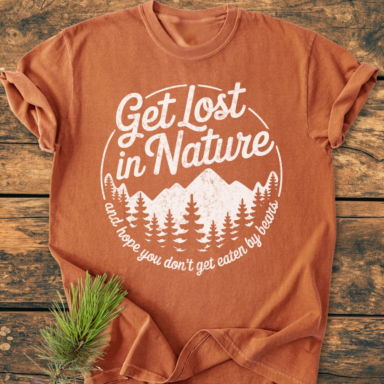 Get Lost In Nature - Vintage Wash Tee with mountain and pine tree graphic, displayed on wood. Accompanied by a pine sprig, emphasizing its nature-inspired theme and relaxed, cozy fit.