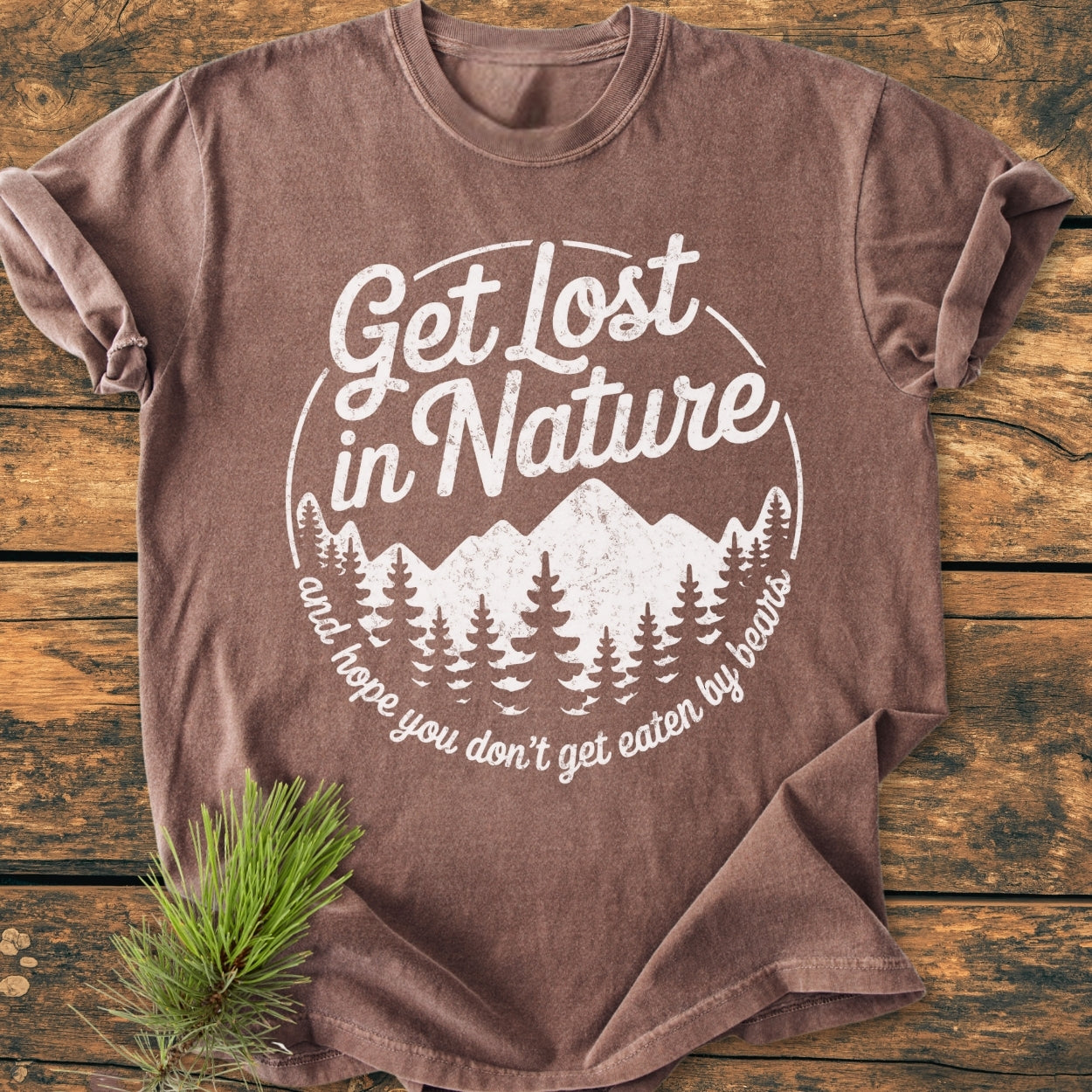 Get Lost In Nature - Vintage Wash Tee features a circular graphic with pine trees and mountains, and a playful slogan, laid flat on a rustic surface with a pine sprig detail.