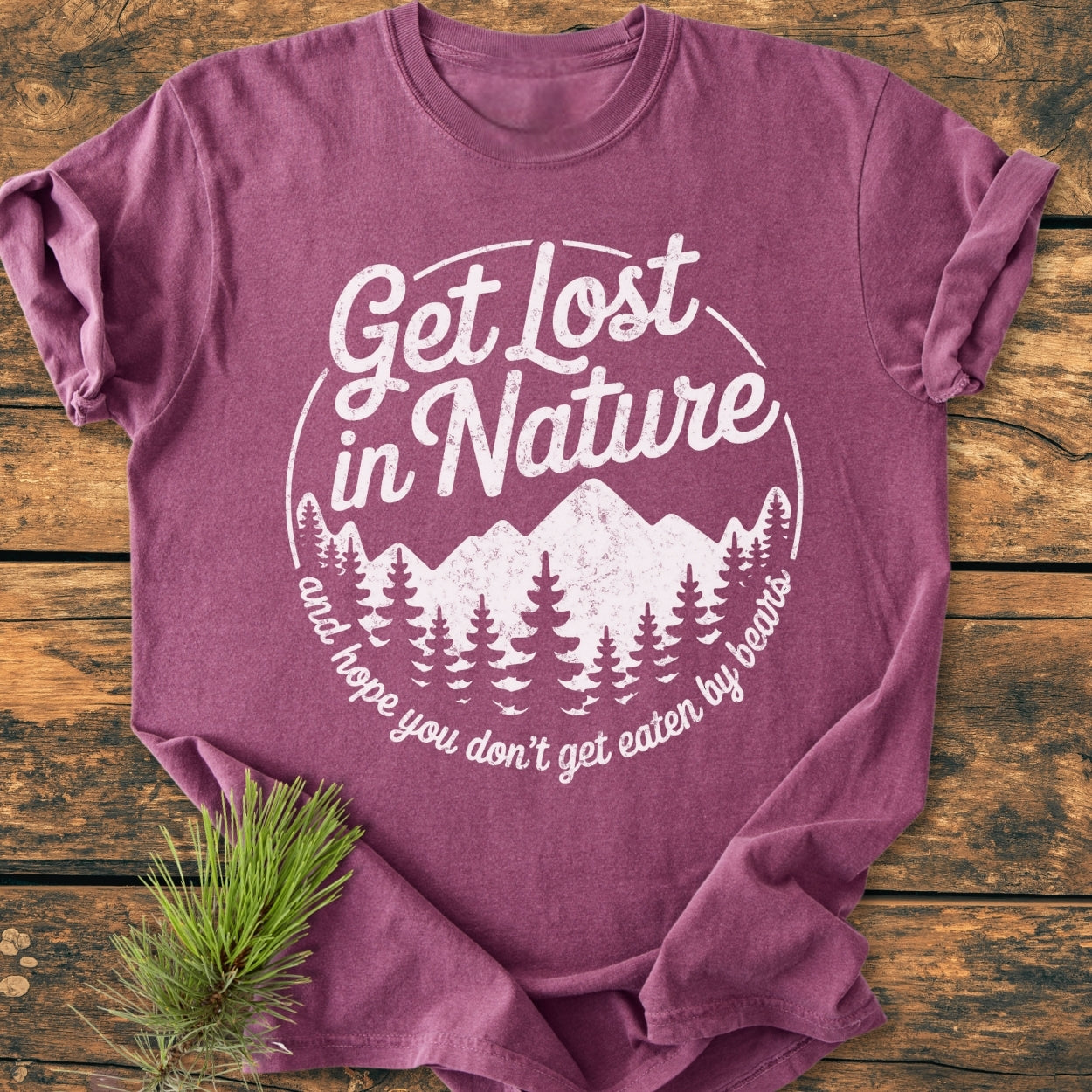 Get Lost In Nature - Vintage Wash Tee featuring a white graphic of pine trees and mountains, with playful text on a soft, relaxed fit, heather-purple T-shirt, laid on a wooden surface.