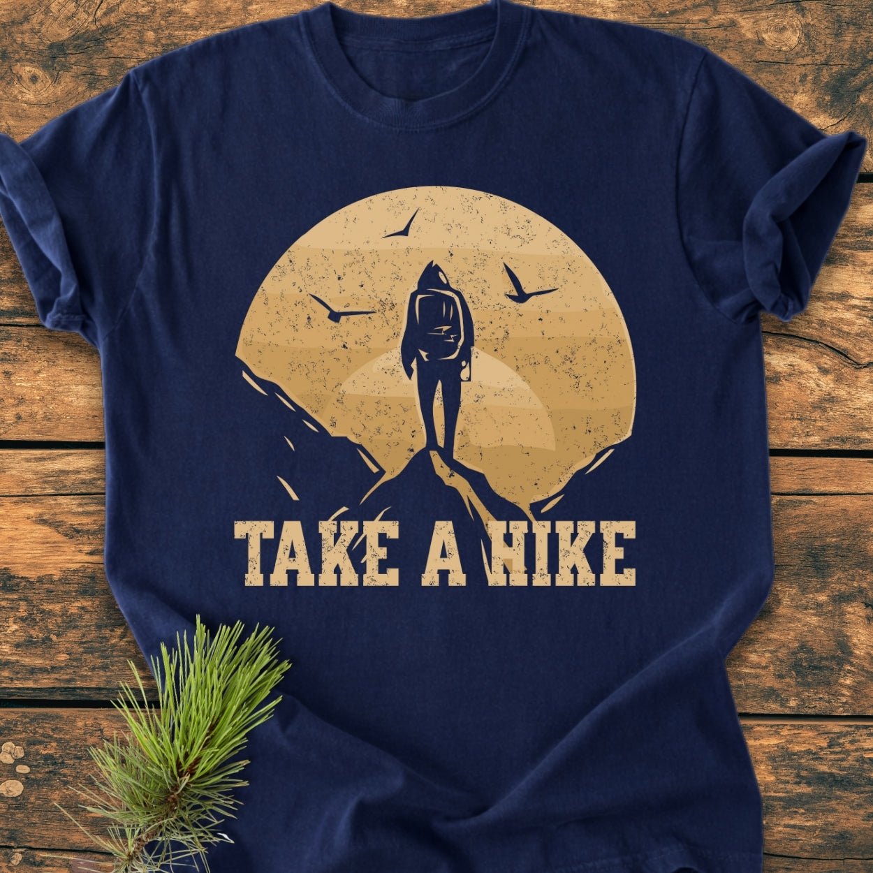 Take A Hike - Vintage Wash Tee, featuring a hiker silhouette with a sun backdrop and flying birds, laid on rustic wood. Includes a pine sprig, showcasing its relaxed fit and durable design.