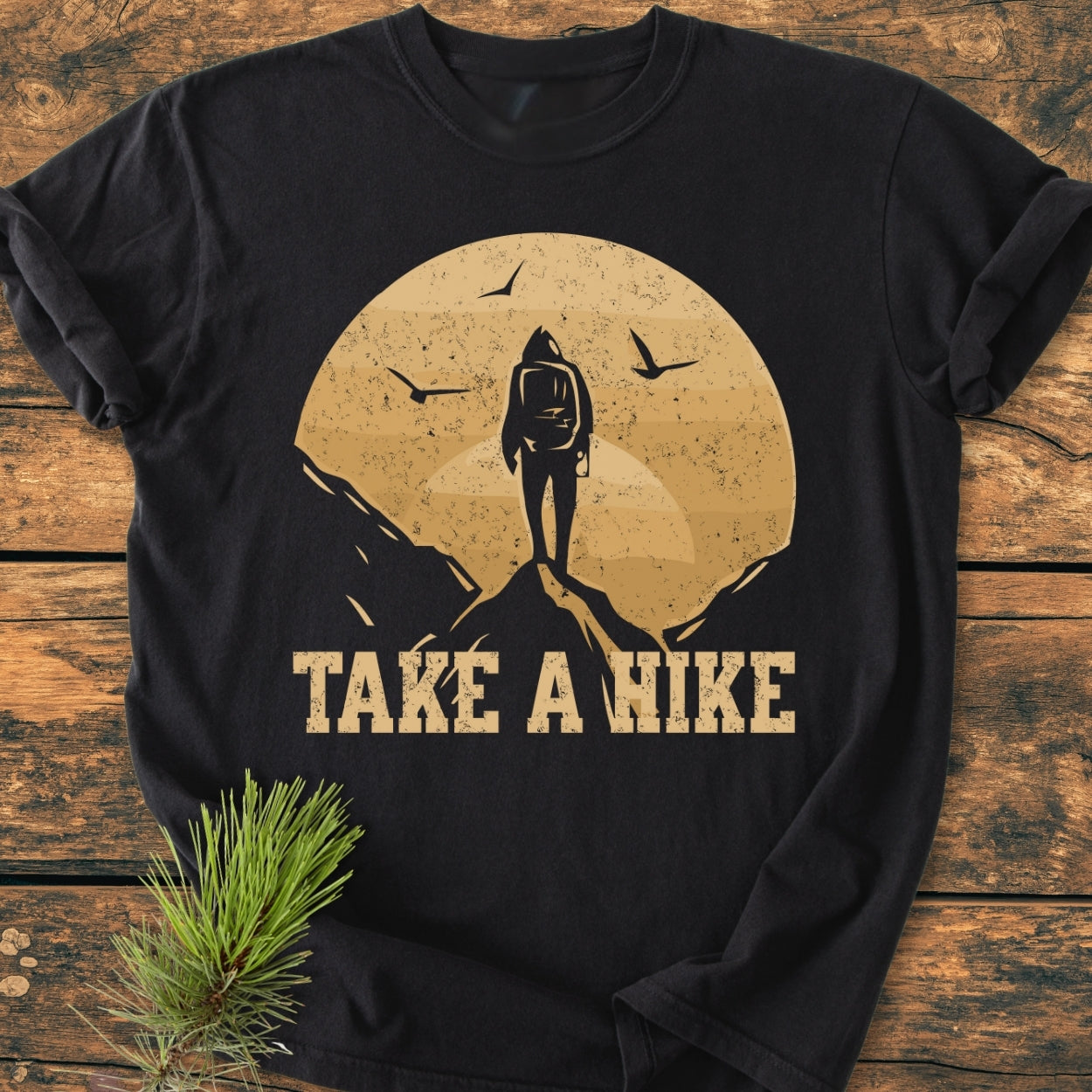 Take A Hike - Vintage Wash Tee features a distressed graphic of a backpacker on a peak with a circular backdrop and birds, laid flat with a pine sprig accentuating its outdoorsy theme.