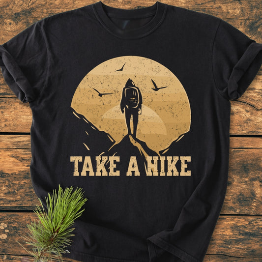 Take A Hike - Vintage Wash Tee features a distressed graphic of a backpacker on a peak with a circular backdrop and birds, laid flat with a pine sprig accentuating its outdoorsy theme.