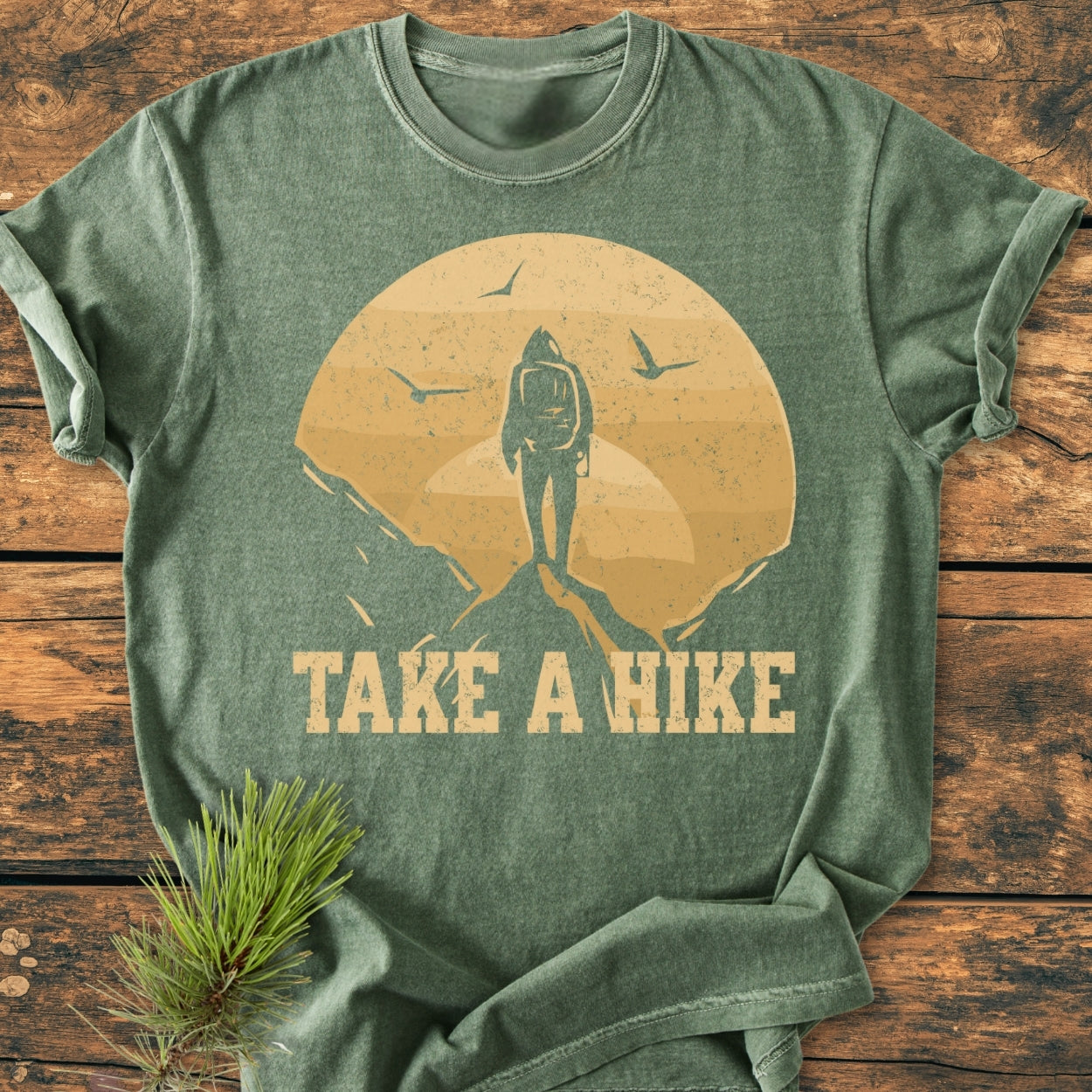 Take A Hike - Vintage Wash Tee featuring a lone hiker graphic with a setting sun and birds, laid on wood with pine sprig. Soft, durable, 100% ring-spun cotton in relaxed fit.
