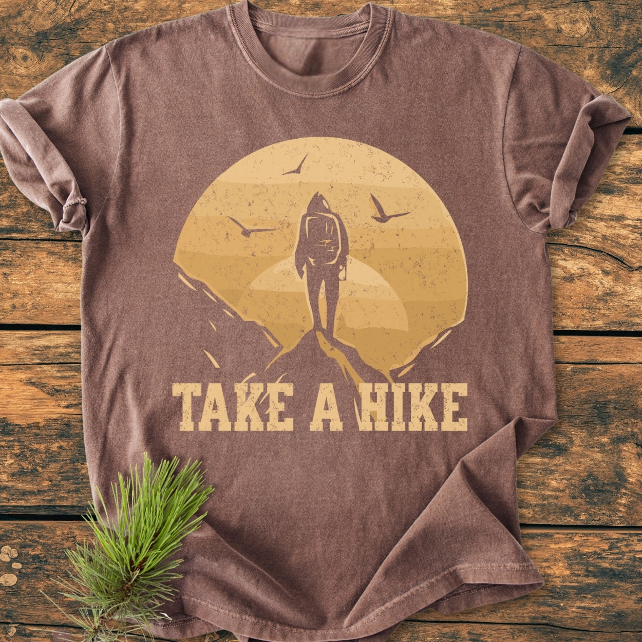 Take A Hike - Vintage Wash Tee: A brown, short-sleeve T-shirt featuring a hiker graphic with setting sun and slogan TAKE A HIKE, laid on wood with pine sprig, highlighting the tee's rugged outdoor theme.