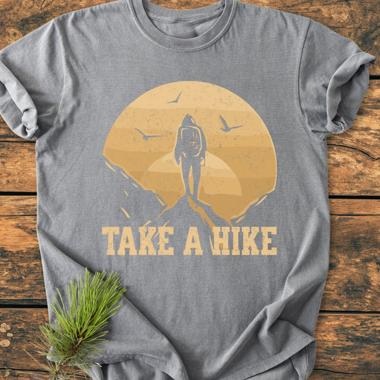 Take A Hike - Vintage Wash Tee featuring a backpacker silhouette with a sun and birds graphic, laid on wood. Made from garment-dyed, soft-washed cotton, showcasing relaxed fit and durability.