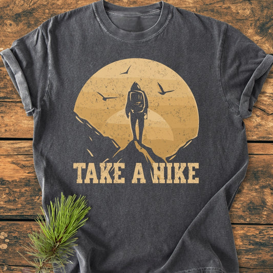 Take A Hike - Vintage Wash Tee: Dark-gray tee features a hiker graphic with sun/moon and birds, rugged TAKE A HIKE text; soft, relaxed fit, double-stitched, laid on wooden surface with spruce twig.