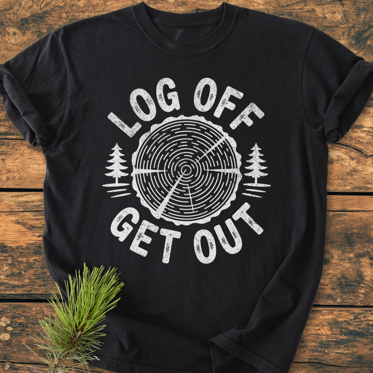 Log Off Get Out - Vintage Wash Tee features a tree-ring graphic with pine silhouettes on a black shirt, laid on wood, capturing an outdoorsy aesthetic with a nearby pine sprig accent.