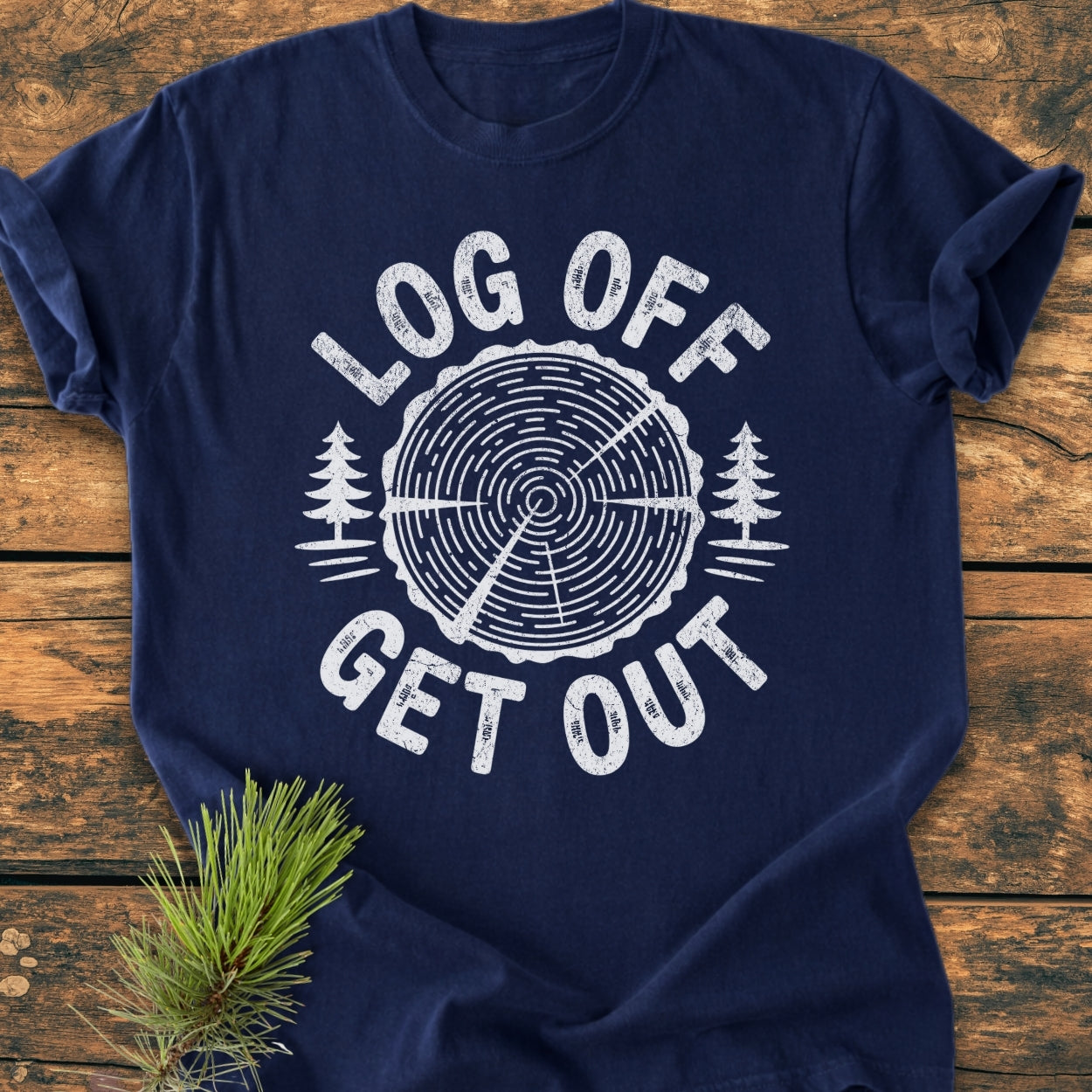 Log Off Get Out - Vintage Wash Tee features a white tree-ring graphic with pine trees and slogan on a navy-blue Comfort Colors T-shirt, laid on wooden planks with a pine branch nearby.