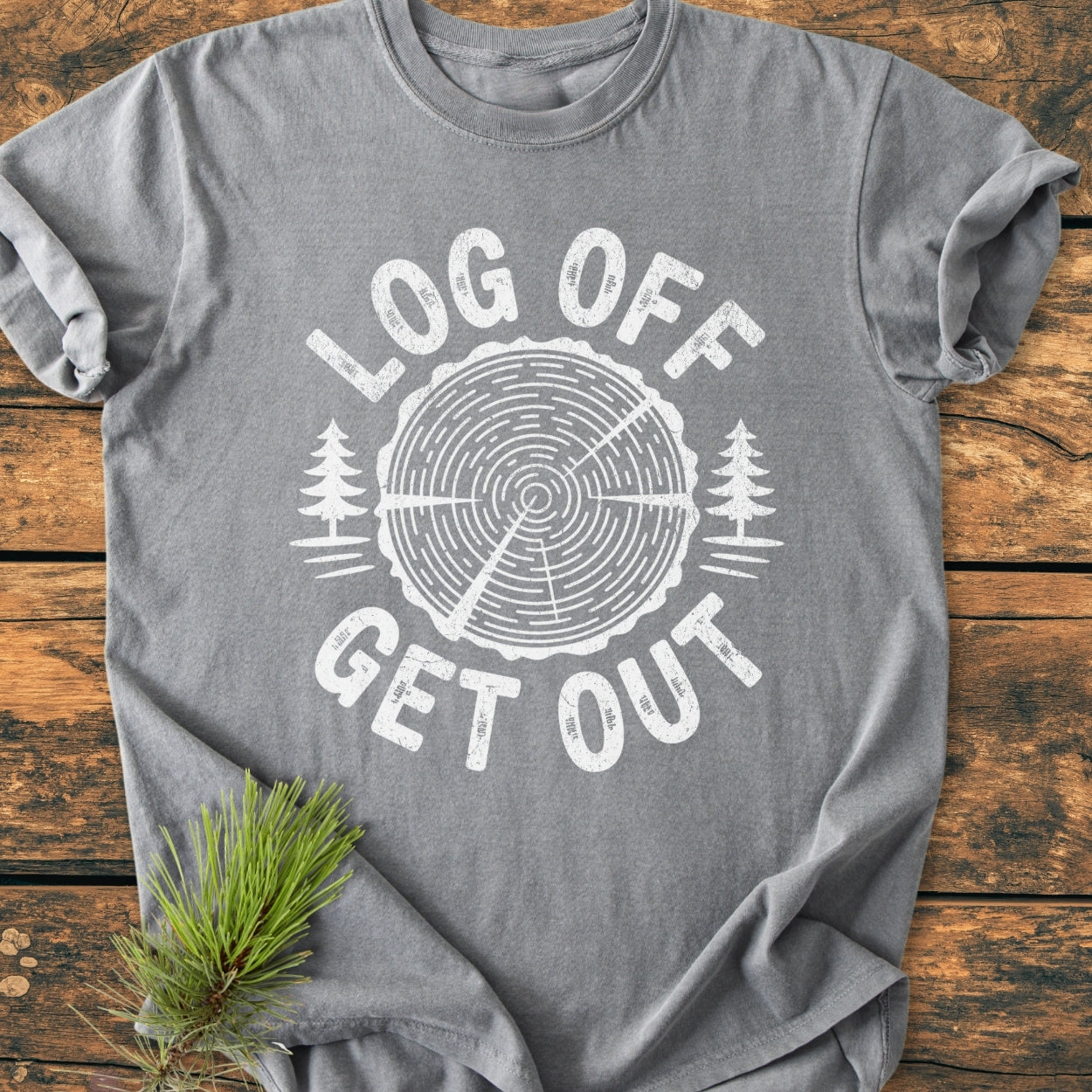 Log Off Get Out - Vintage Wash Tee: Heather-gray shirt with LOG OFF and GET OUT text around a log graphic, featuring pine trees. Laid flat, showcasing relaxed fit and pine bough accent.