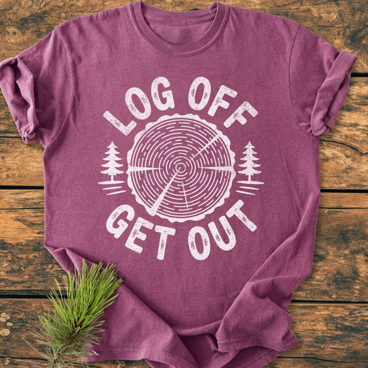 Log Off Get Out - Vintage Wash Tee features a tree trunk graphic with pine trees and text on a maroon-purple short-sleeve tee, laid flat with a pine sprig accent.