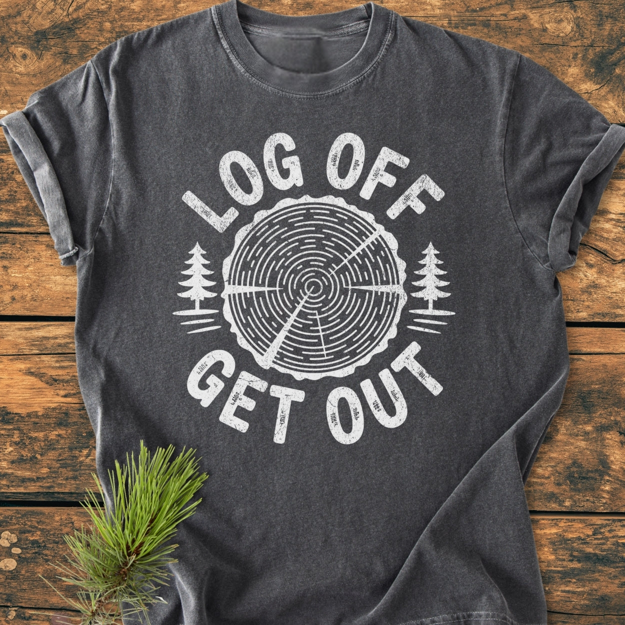 Log Off Get Out - Vintage Wash Tee: Dark-gray short-sleeve T-shirt featuring a tree-ring design with LOG OFF and GET OUT text, laid flat on wood, pine sprig accentuating its outdoorsy appeal.