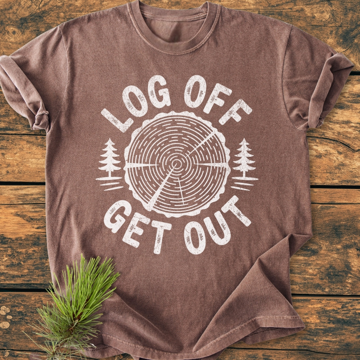 Log Off Get Out - Vintage Wash Tee: brown, short-sleeved with a stylized tree-ring design and text. Laid flat on wood, showcasing its relaxed fit and durable, soft garment-dyed fabric.
