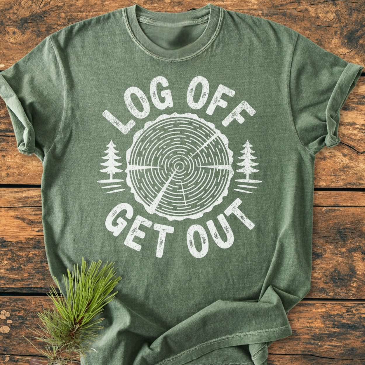 Heather-green Log Off Get Out - Vintage Wash Tee featuring a tree trunk graphic and slogan, laid flat with a pine branch beside it on a wooden surface, showcasing its relaxed fit.