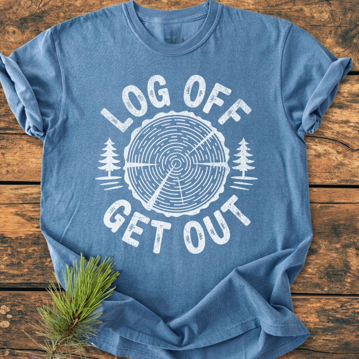 Heather-blue Log Off Get Out - Vintage Wash Tee features a white tree trunk design with pine trees and slogan, laid flat on wood, embodying an outdoors theme. Pine sprig accentuates nature-inspired style.