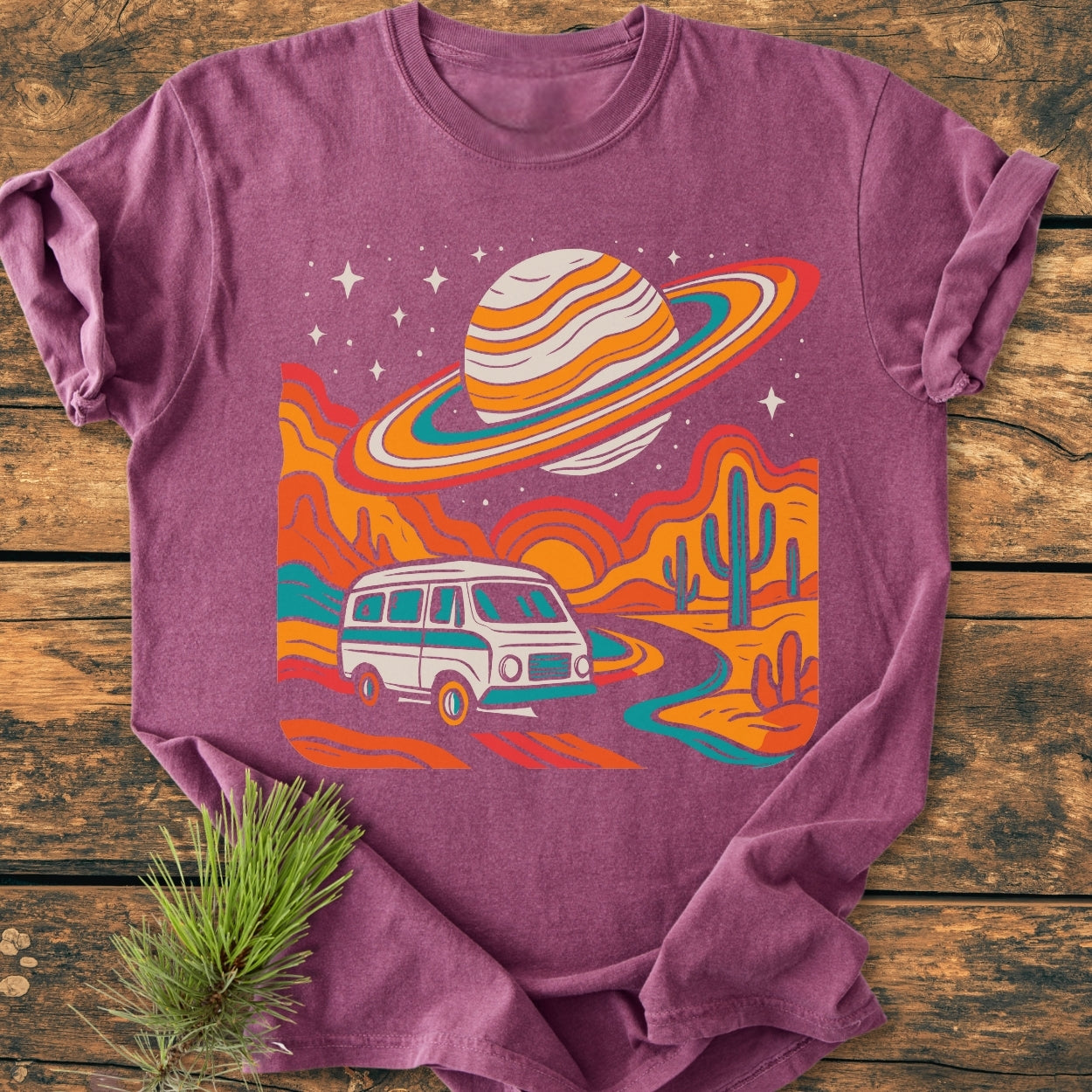 Psychedelic Van Journey - Vintage Wash Tee features a retro camper van on a desert road under a giant ringed planet, all in vibrant colors, laid on rustic wood with pine sprig.