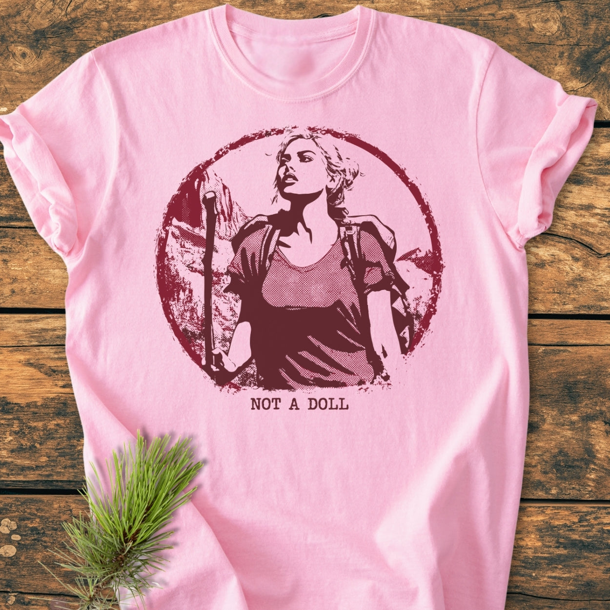 Not A Doll - Vintage Wash Tee features a burgundy graphic of a woman hiking, with NOT A DOLL text below, on a soft, pale-pink, 100% ring-spun cotton fabric, laid on rustic wood.