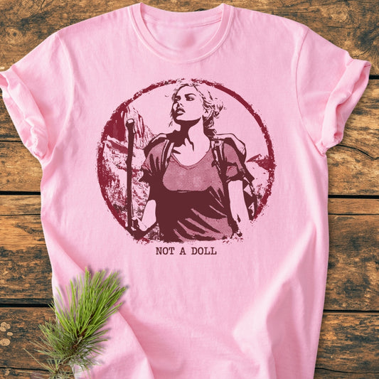 Not A Doll - Vintage Wash Tee features a burgundy graphic of a woman hiking, with NOT A DOLL text below, on a soft, pale-pink, 100% ring-spun cotton fabric, laid on rustic wood.