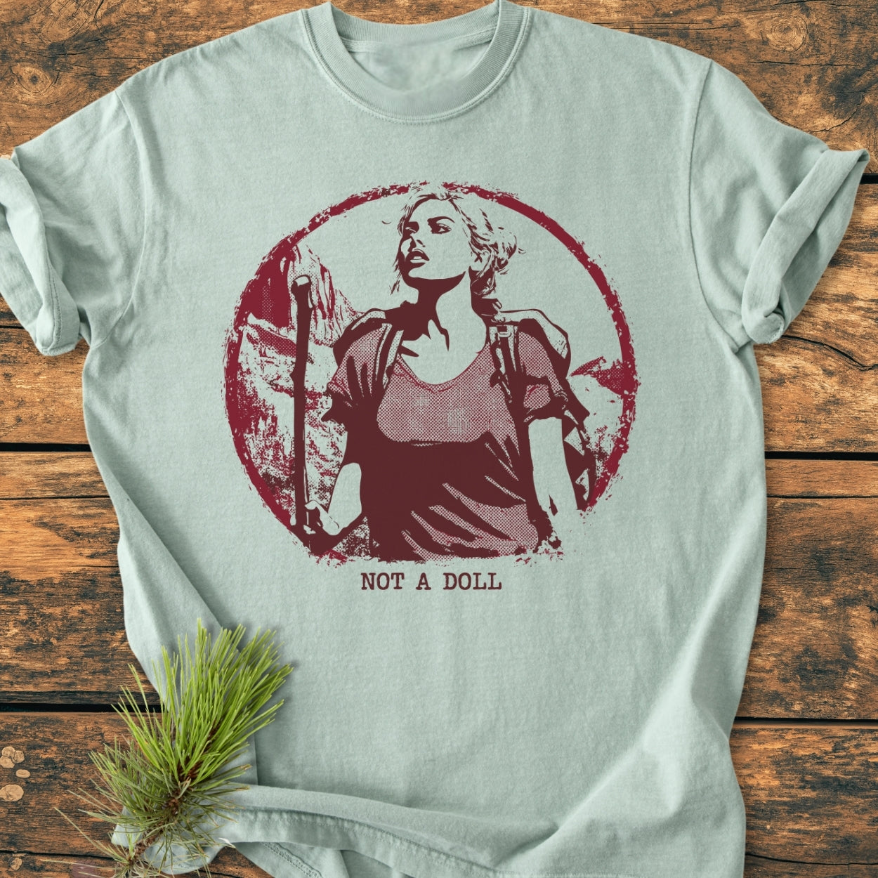 Not A Doll - Vintage Wash Tee features a circular graphic of a hiker against mountains in distressed dark-red ink, laid flat with a pine sprig accent. Made from soft, durable, pre-shrunk cotton.