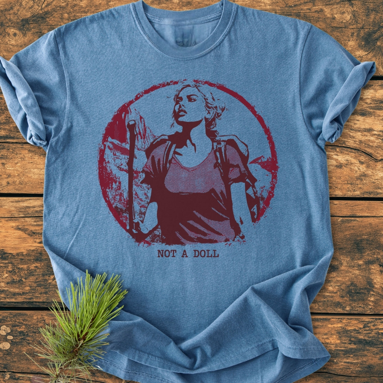 Heather-blue Not A Doll - Vintage Wash Tee with a central graphic of a backpacking woman, laid flat on wood. Screen-printed message “NOT A DOLL.” emphasizes empowerment, enhancing its relaxed, durable design.