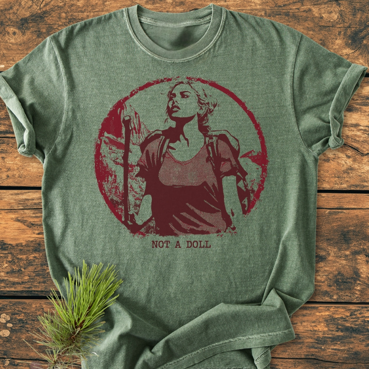 Not A Doll - Vintage Wash Tee in heather-olive features a maroon vignette of a hiking woman and pine bough accent, showcasing a durable, relaxed fit with soft, garment-dyed cotton.