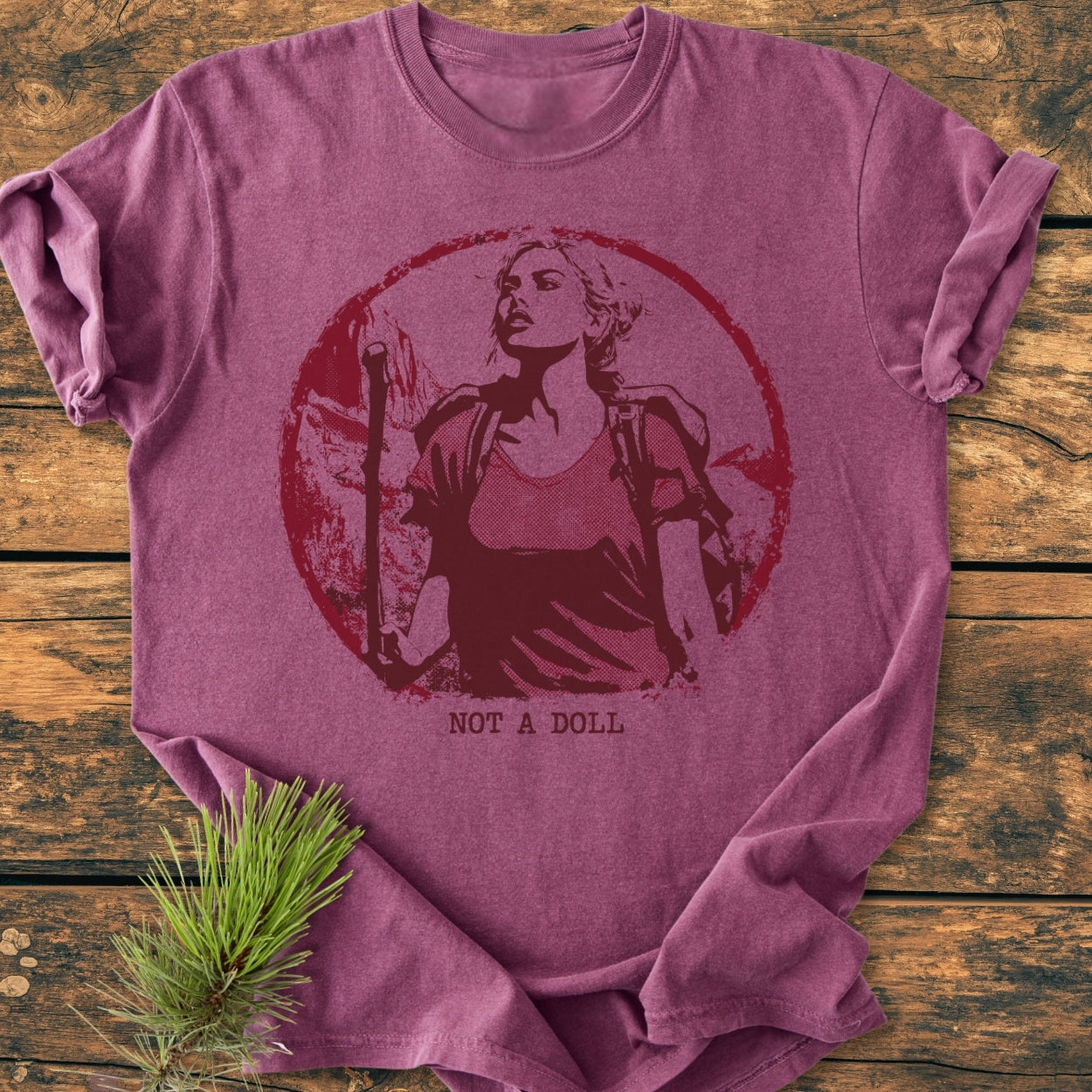 Not A Doll - Vintage Wash Tee featuring a heather-purple color, circular graphic of a determined woman with hiking gear, and NOT A DOLL text, displayed on a wooden surface with pine sprig.