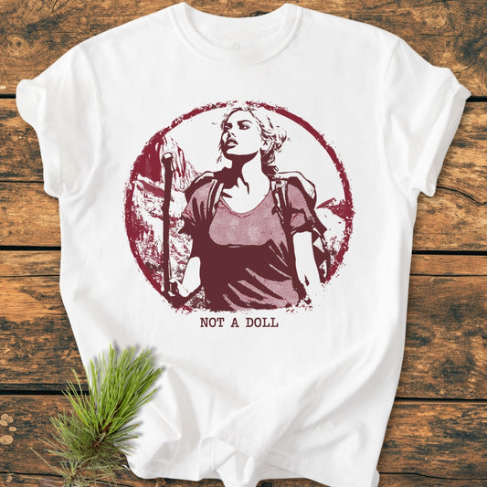 Not A Doll - Vintage Wash Tee displayed on wood, featuring a circular graphic of a woman hiking with a backpack. Beside the shirt, a pine sprig adds a natural touch.