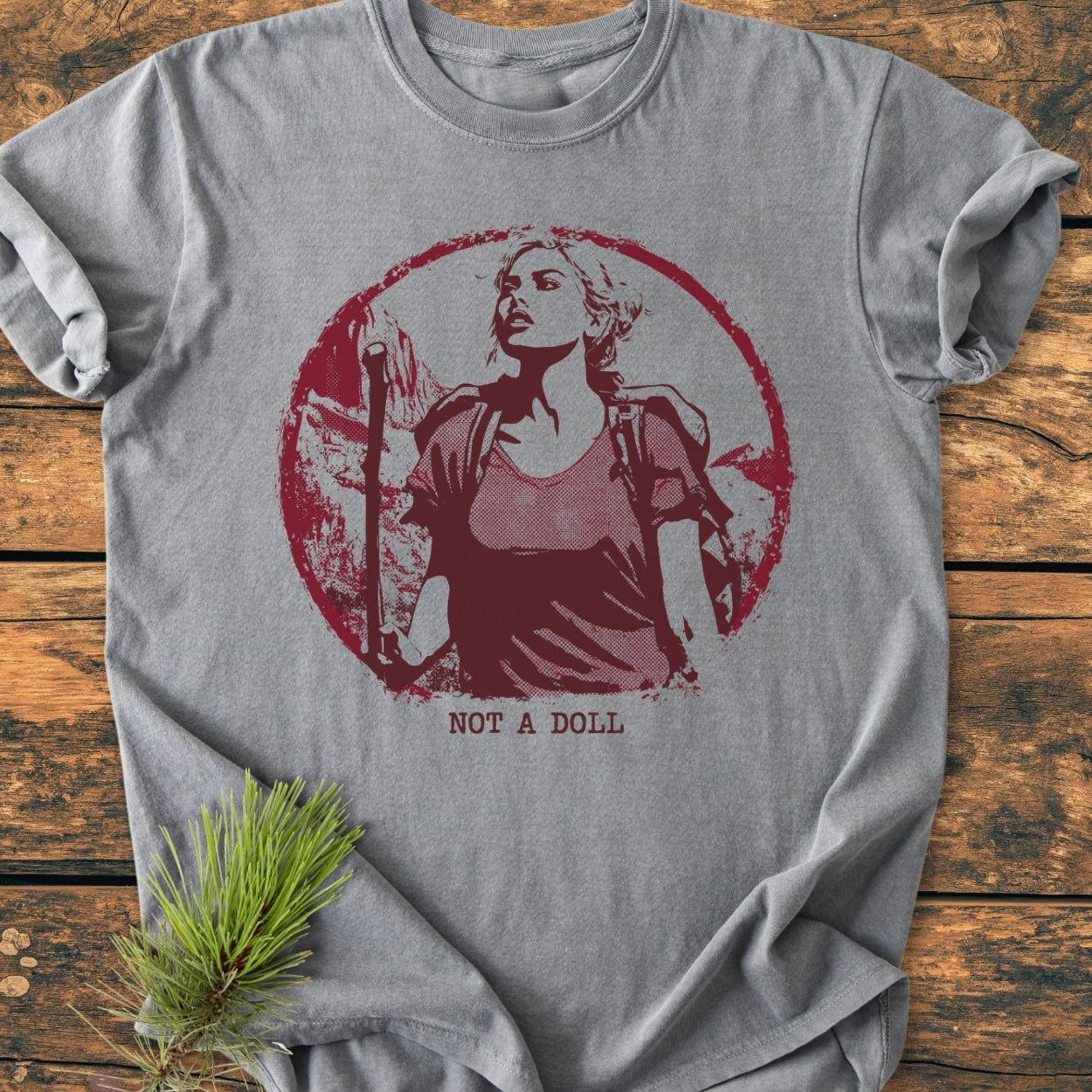 Not A Doll - Vintage Wash Tee: Heather-gray T-shirt with a red distressed graphic of a woman with a backpack and hiking pole, styled with a pine sprig on a wooden surface.