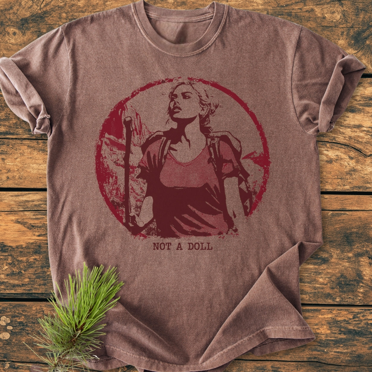 Not A Doll - Vintage Wash Tee features a distressed graphic of a woman with a backpack and hiking staff, centered on a soft, brown T-shirt, laid flat on a wood surface.