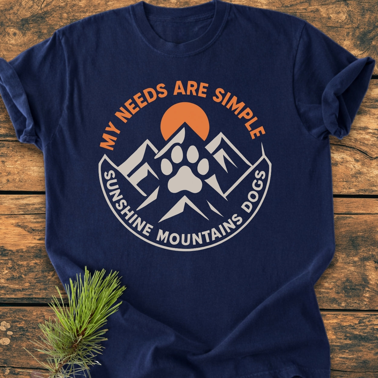 Simple Needs. Mountains, Sunshine & Dog - Vintage Wash Tee featuring a circular mountain scene with a dog-paw silhouette, orange sun, and slogan, laid on a wooden surface with a pine sprig.