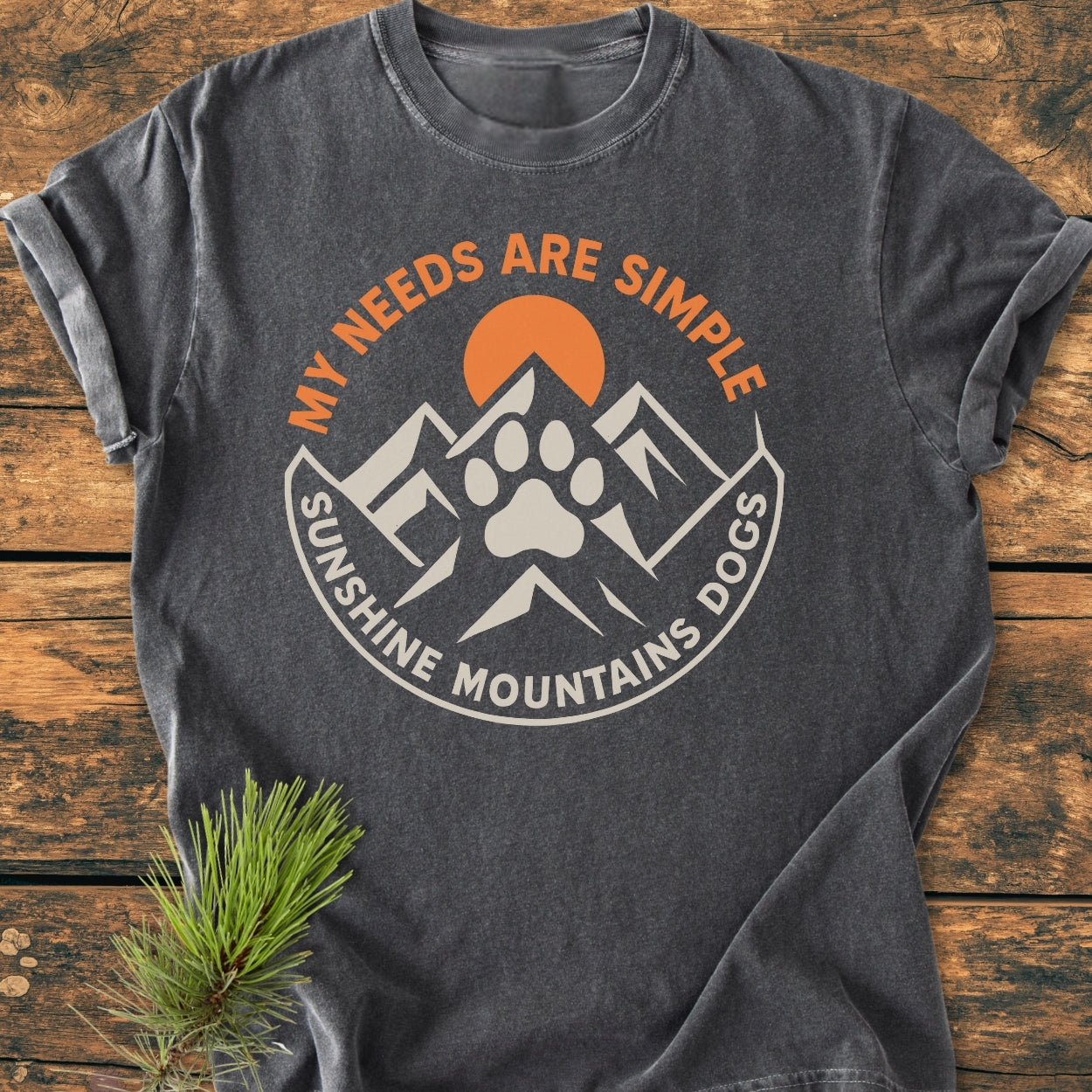 Charcoal-gray Simple Needs. Mountains, Sunshine & Dog - Vintage Wash Tee with a circular badge graphic featuring an orange sun, cream mountains, and a dog paw print on a wooden surface.