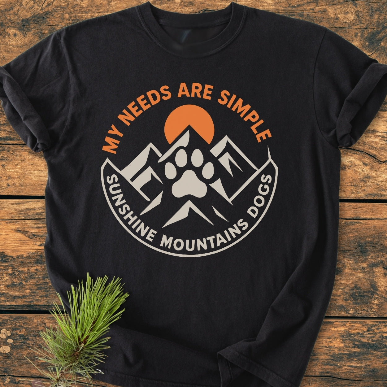 Simple Needs. Mountains, Sunshine & Dog - Vintage Wash Tee featuring stylized mountains, an orange sun, and a dog paw graphic, printed on a black Comfort Colors garment-dyed T-shirt laid on wood surface.