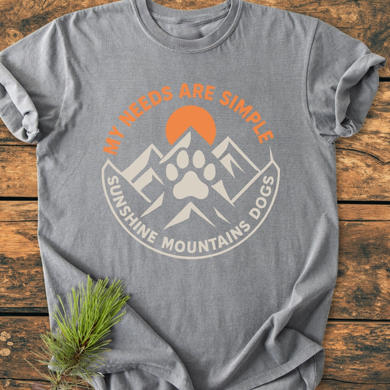 Simple Needs. Mountains, Sunshine & Dog - Vintage Wash Tee in heather-gray, featuring a mountain and paw print design with a rising sun. Laid flat with a pine sprig accent.