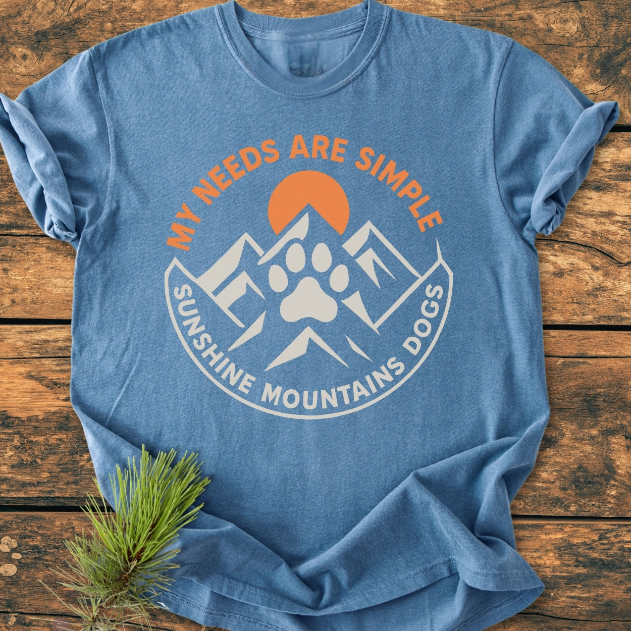 Simple Needs. Mountains, Sunshine & Dog - Vintage Wash Tee in blue, featuring an outdoor-themed graphic with a sun, mountain peaks, and paw print. Laid flat with a pine branch accent.