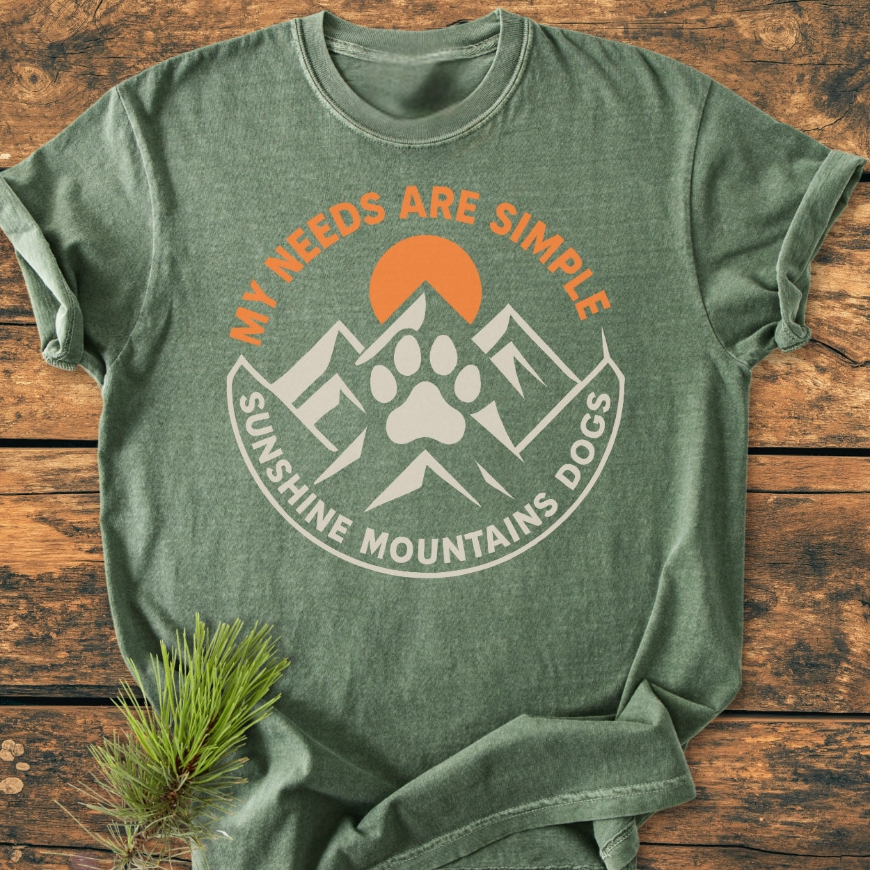 Simple Needs. Mountains, Sunshine & Dog - Vintage Wash Tee: Heather-green T-shirt featuring a mountain and sun graphic with a dog-paw silhouette, laid on a wooden surface with a pine sprig beside it.