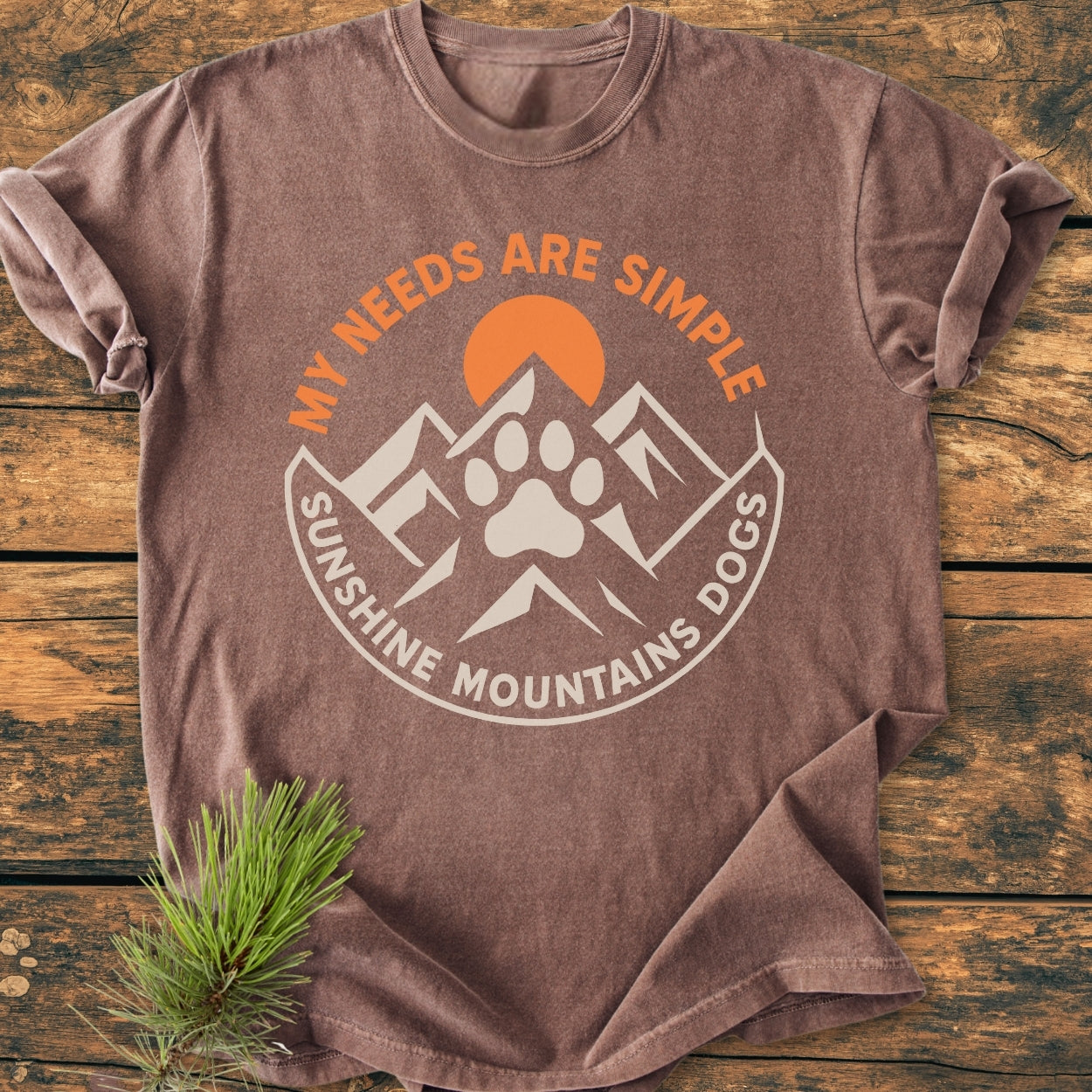 Simple Needs. Mountains, Sunshine & Dog - Vintage Wash Tee featuring a circular design with mountains, sun, and paw print, laid on wood. Includes MY NEEDS ARE SIMPLE and SUNSHINE MOUNTAINS DOGS text.