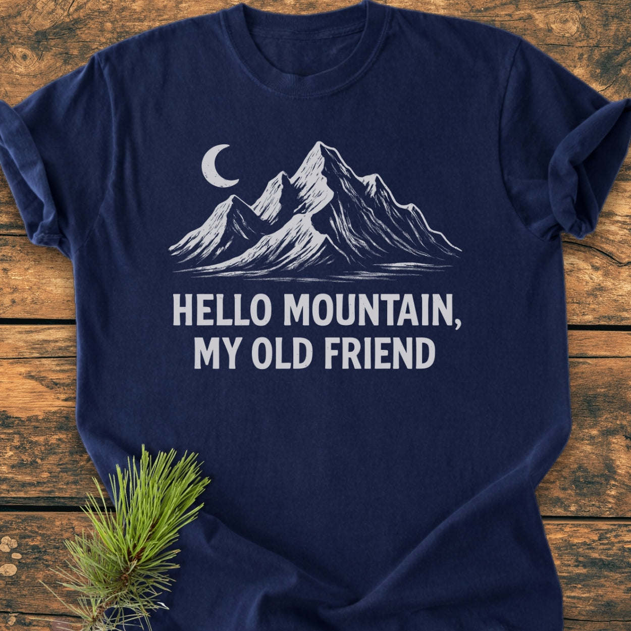 Hello Mountain My Old Friend - Vintage Wash Tee features a white mountain and moon design on a navy-blue T-shirt, laid on wooden surface with pine bough accentuating its rustic appeal.