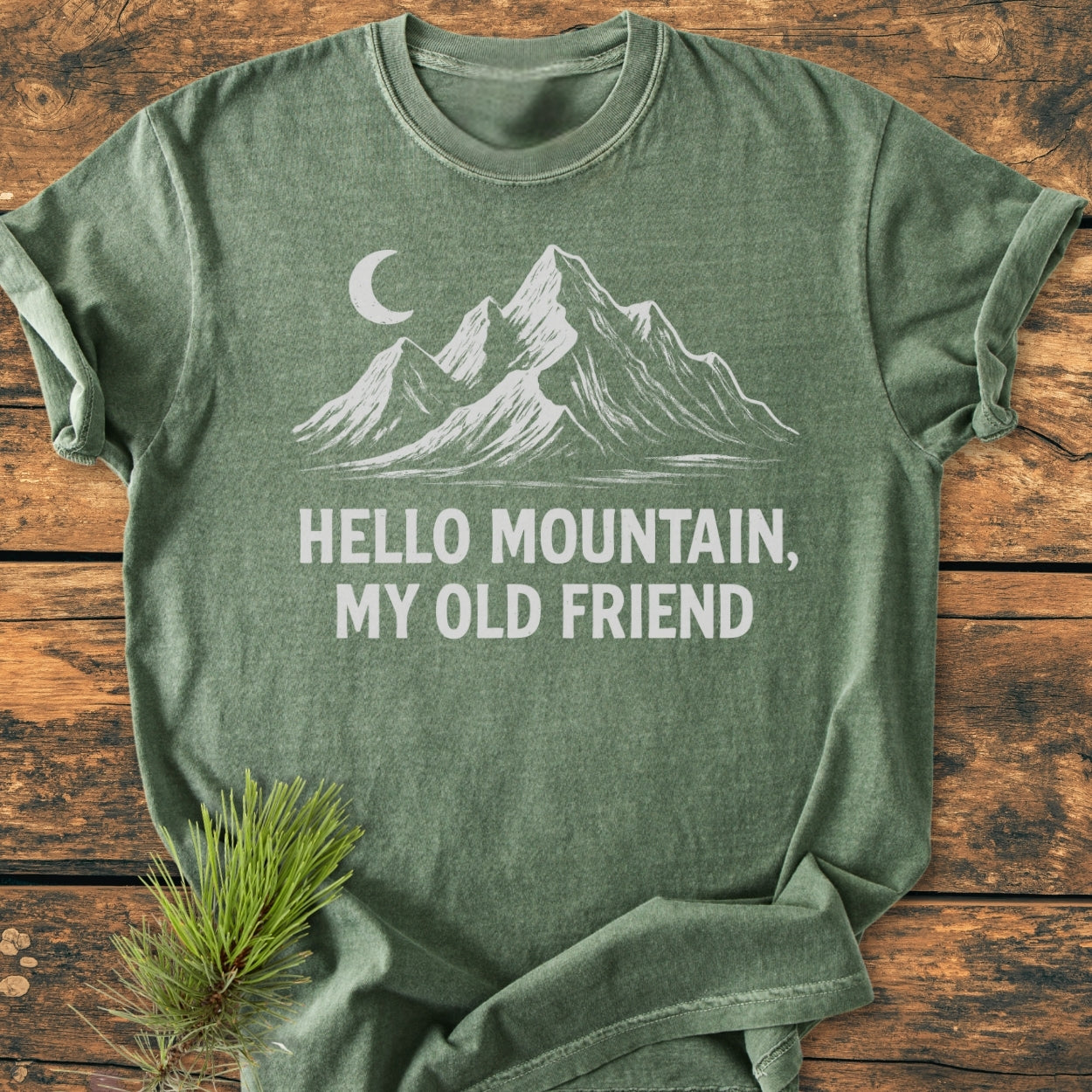 Hello Mountain My Old Friend - Vintage Wash Tee features a forest-green shirt with a white mountain design and crescent moon, laid on wood with a pine sprig beside it.