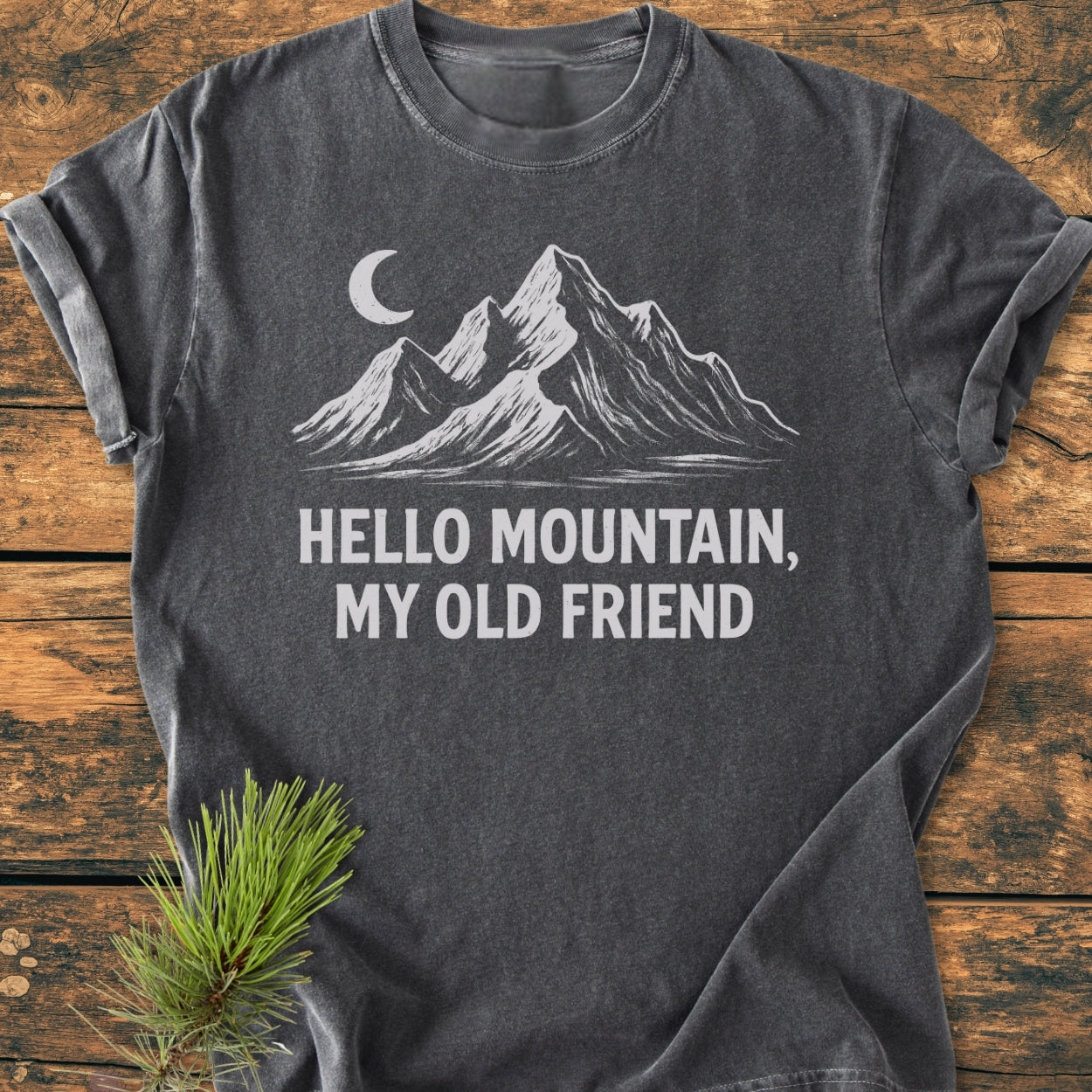Hello Mountain My Old Friend - Vintage Wash Tee features a white mountain and crescent moon graphic on a dark-gray T-shirt, laid on wood with a pine sprig, showcasing its rustic charm.