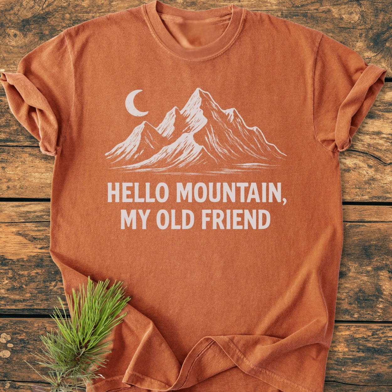 Hello Mountain My Old Friend - Vintage Wash Tee in burnt-orange with mountain and moon print, laid on wood. Features a sprig of pine, highlighting its nature-inspired theme.